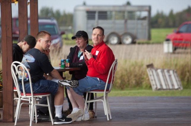 A group of people sit at a table in a park shelter on a summer day. A man in a red sweater is turned in his chair, looking at the camera.