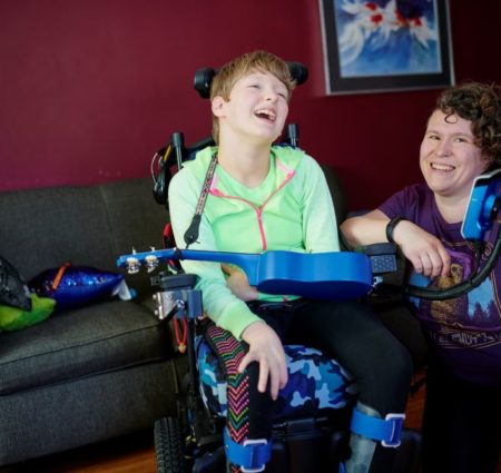 A girl in bright green sits in her wheelchair and laughs. Next to her a woman in purple kneels and smiles at the camera