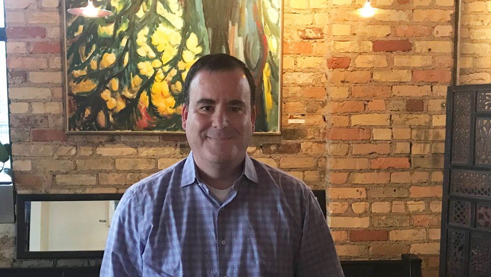 Middle-aged man looks directly at the camera in front of a brick cafe wall.